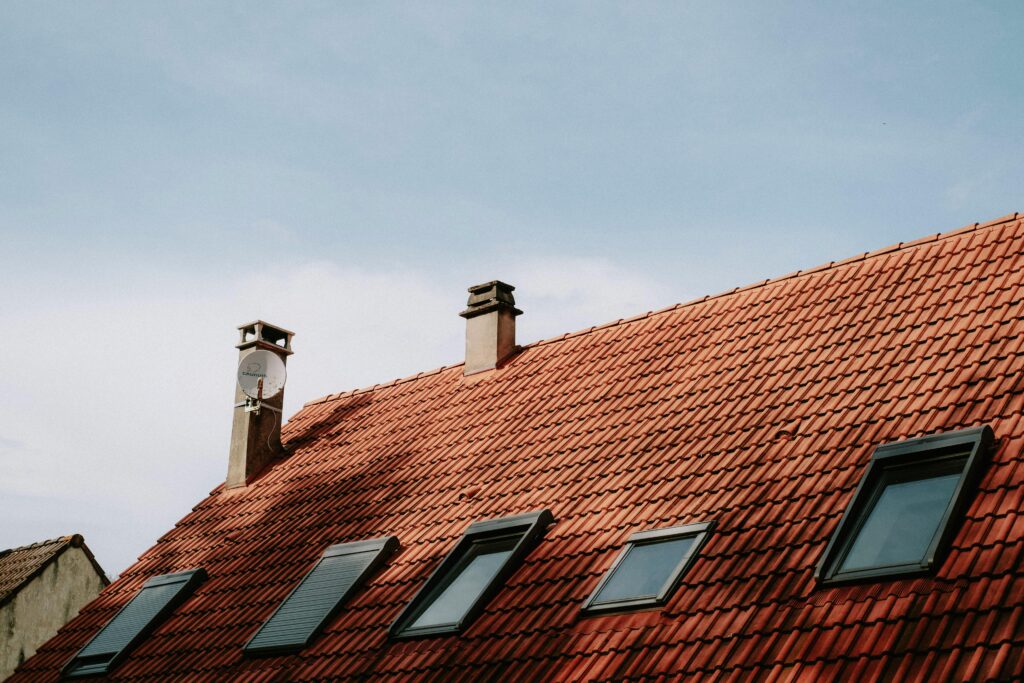 A picturesque red tiled roof featuring chimneys and skylight windows against a blue sky.